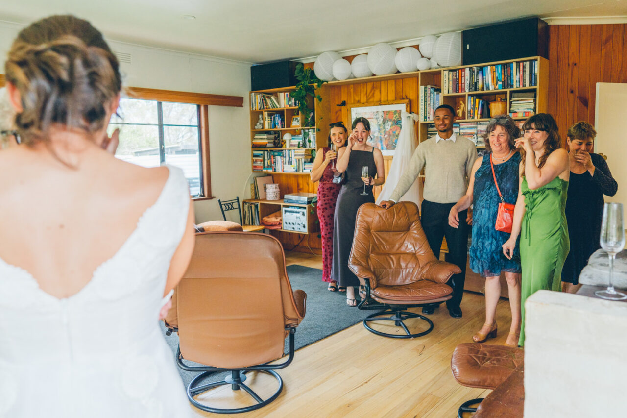 Bride seen from behind in a vintage white wedding dress as friends and family react with tears and laughter seeing her for the first time in the family home