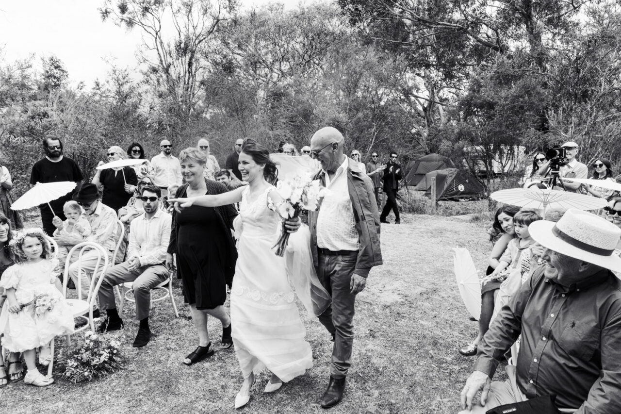 Black and white image of a bride walking down the aisle through a backyard with her parents, guests seated on either side holding parasols