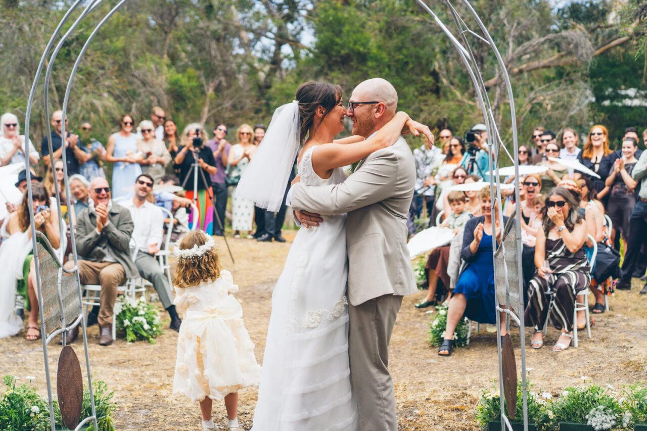 Bride and groom embracing and laughing under a metal arbour after being pronounced married, their young daughter standing in front of them as guests cheer and clap
