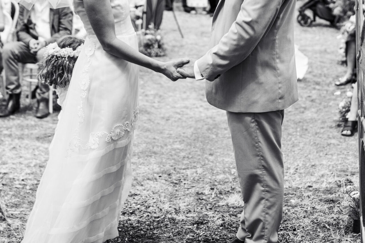 Black and white close up of a bride and groom holding hands during their wedding ceremony, the lace detail of the vintage dress visible