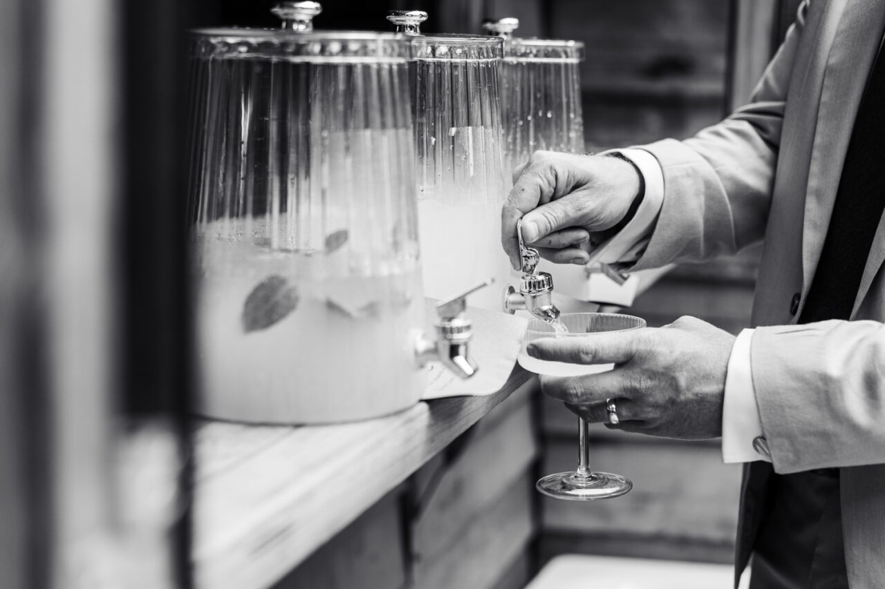 Black and white close up of a person in a suit pouring a cocktail from a glass dispenser at a backyard wedding bar