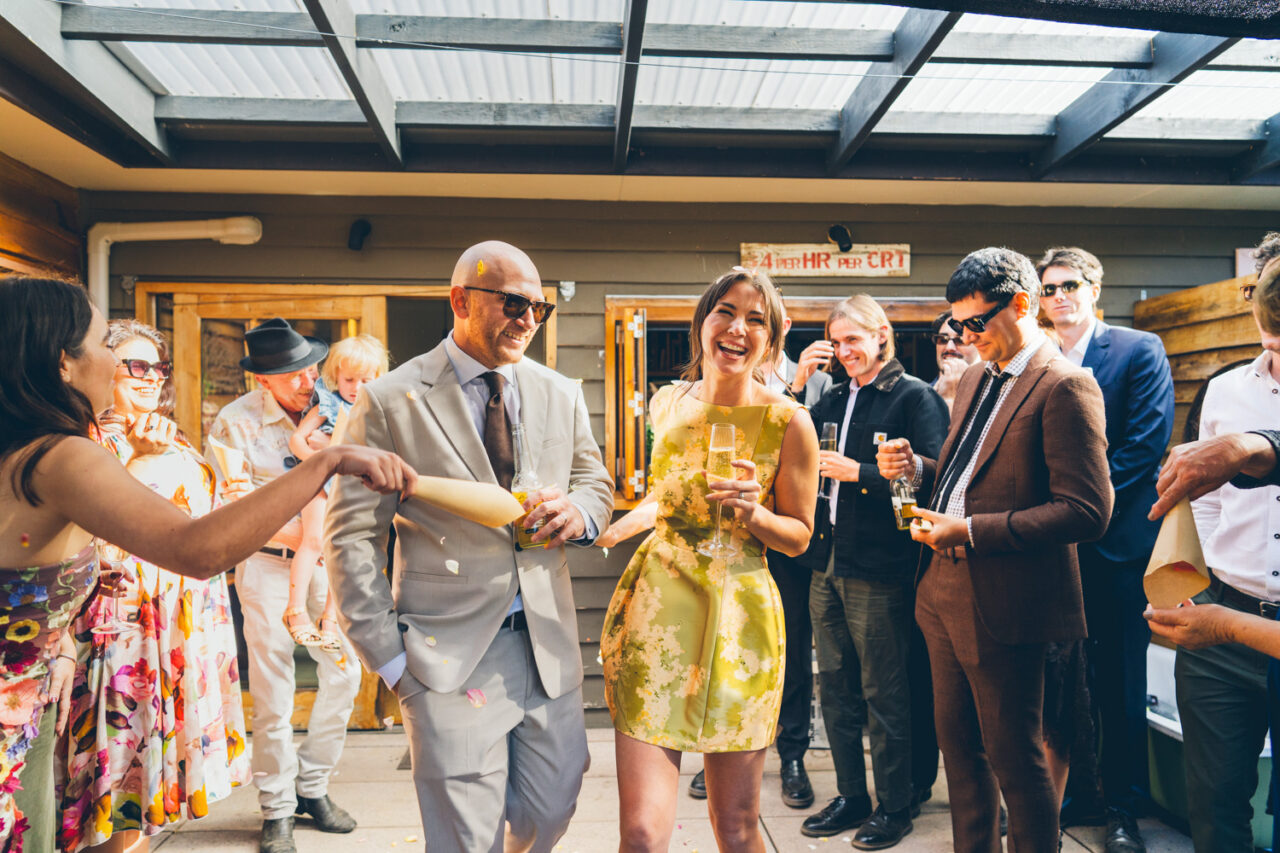 Bride and groom walking through a confetti shower at their backyard wedding reception entrance, surrounded by cheering guests
