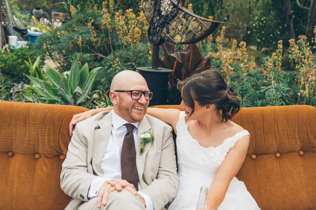 Bride and groom sitting together on a vintage mustard velvet couch in a lush garden, laughing and leaning into each other