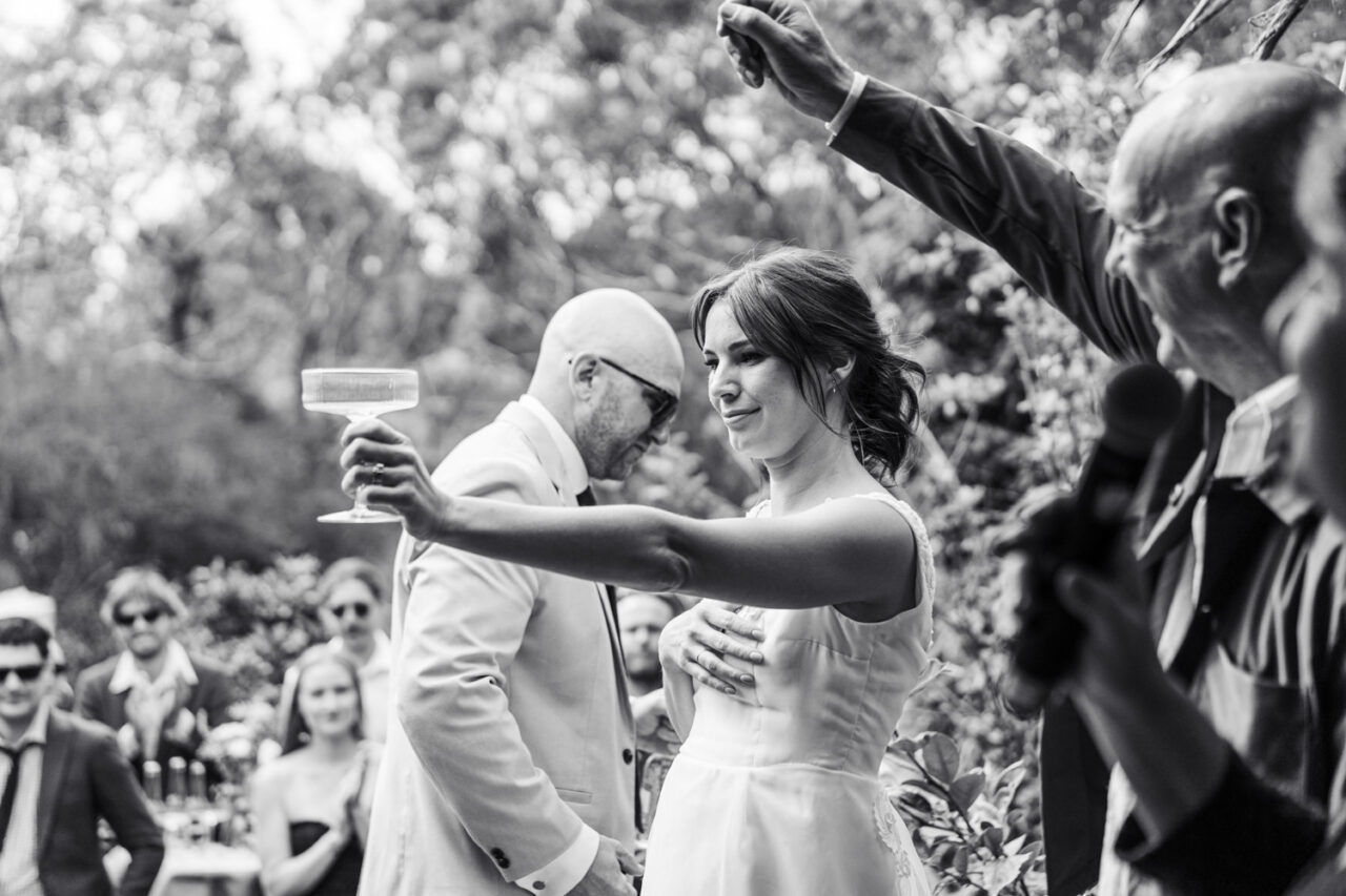 Black and white image of a bride and groom raising their glasses in a toast at their backyard wedding reception surrounded by cheering guests