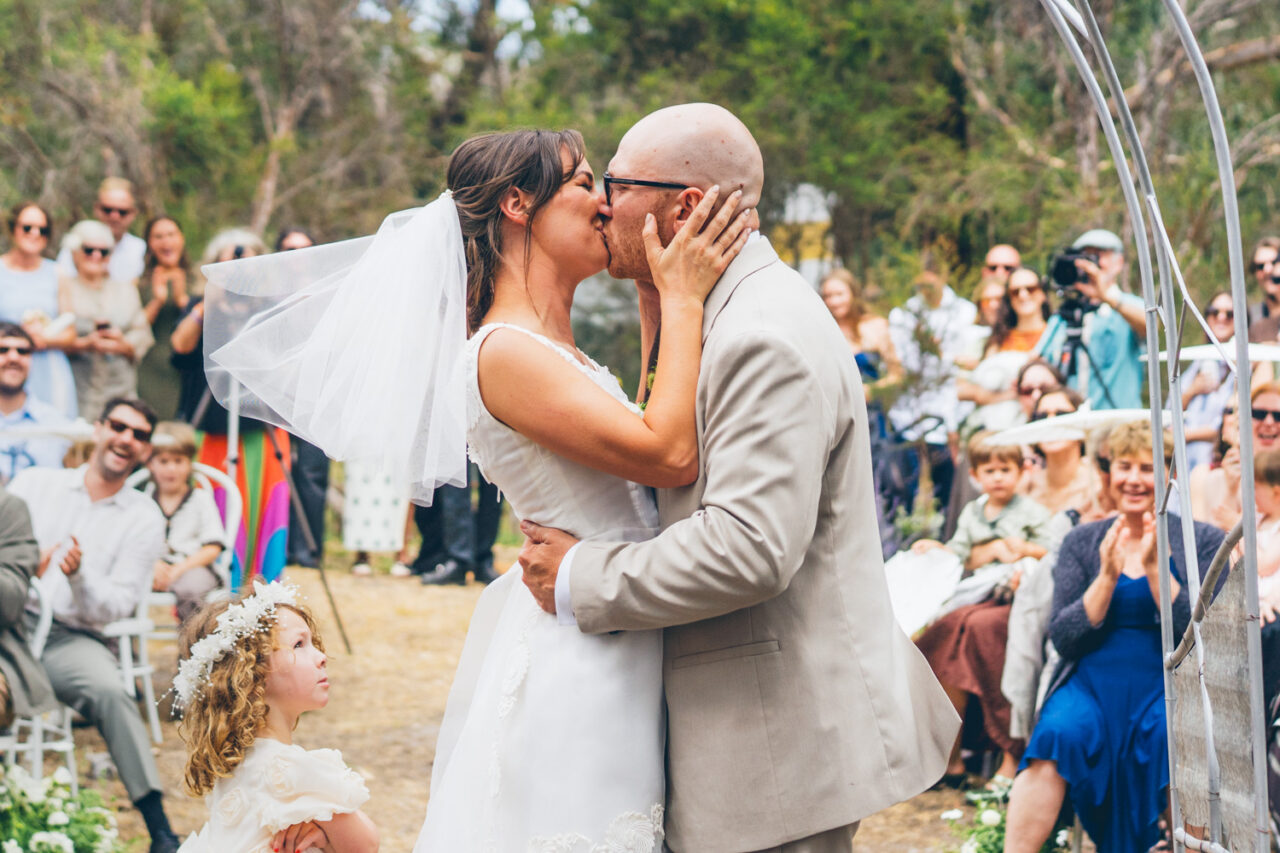 Bride and groom sharing their first kiss under a metal arbour at a backyard wedding on the Surf Coast, their daughter standing below looking up at them