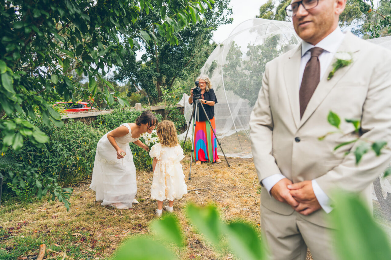 Bride bending down to her young daughter during the first look, while the groom stands in the foreground and the videographer films in the background