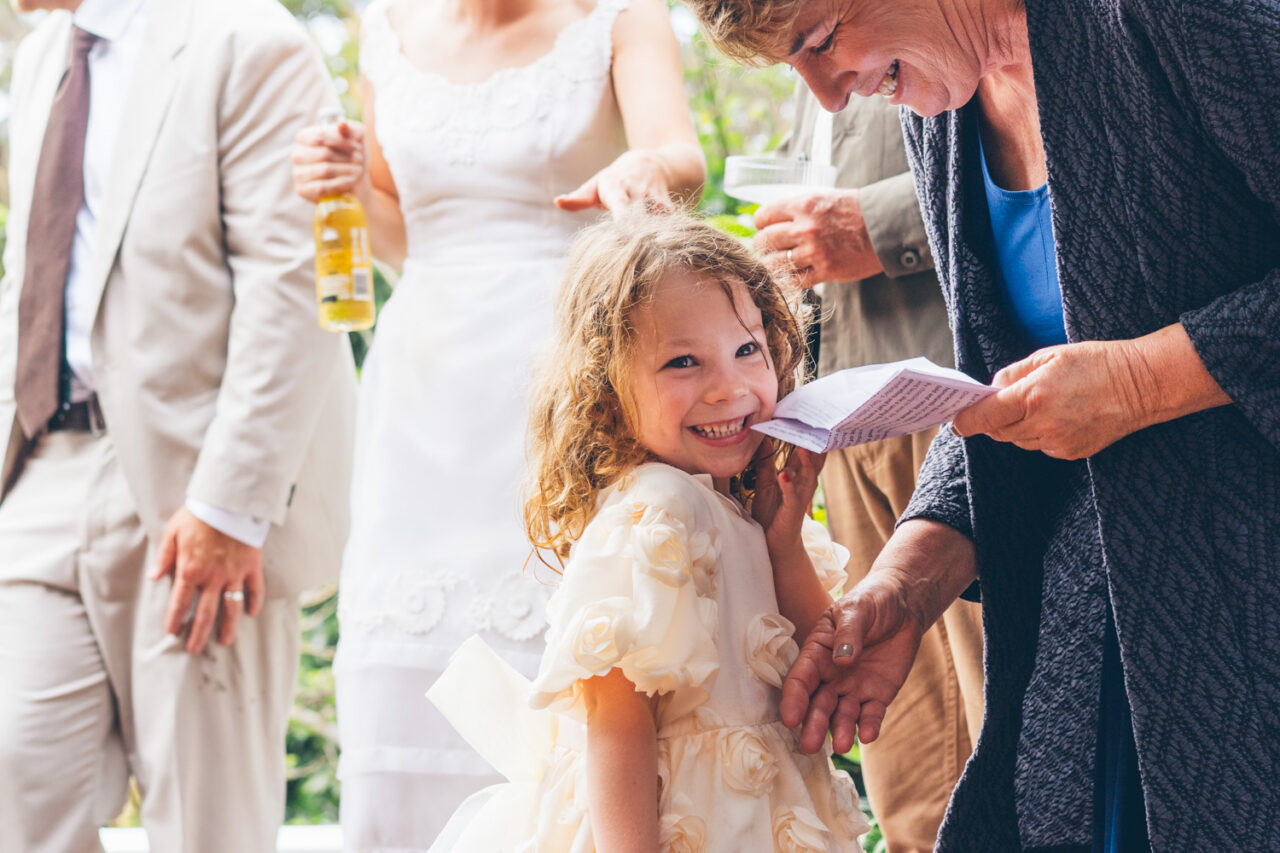Young flower girl in a cream ruffled dress grinning broadly at an older guest holding a ceremony booklet, the bride and groom visible behind them