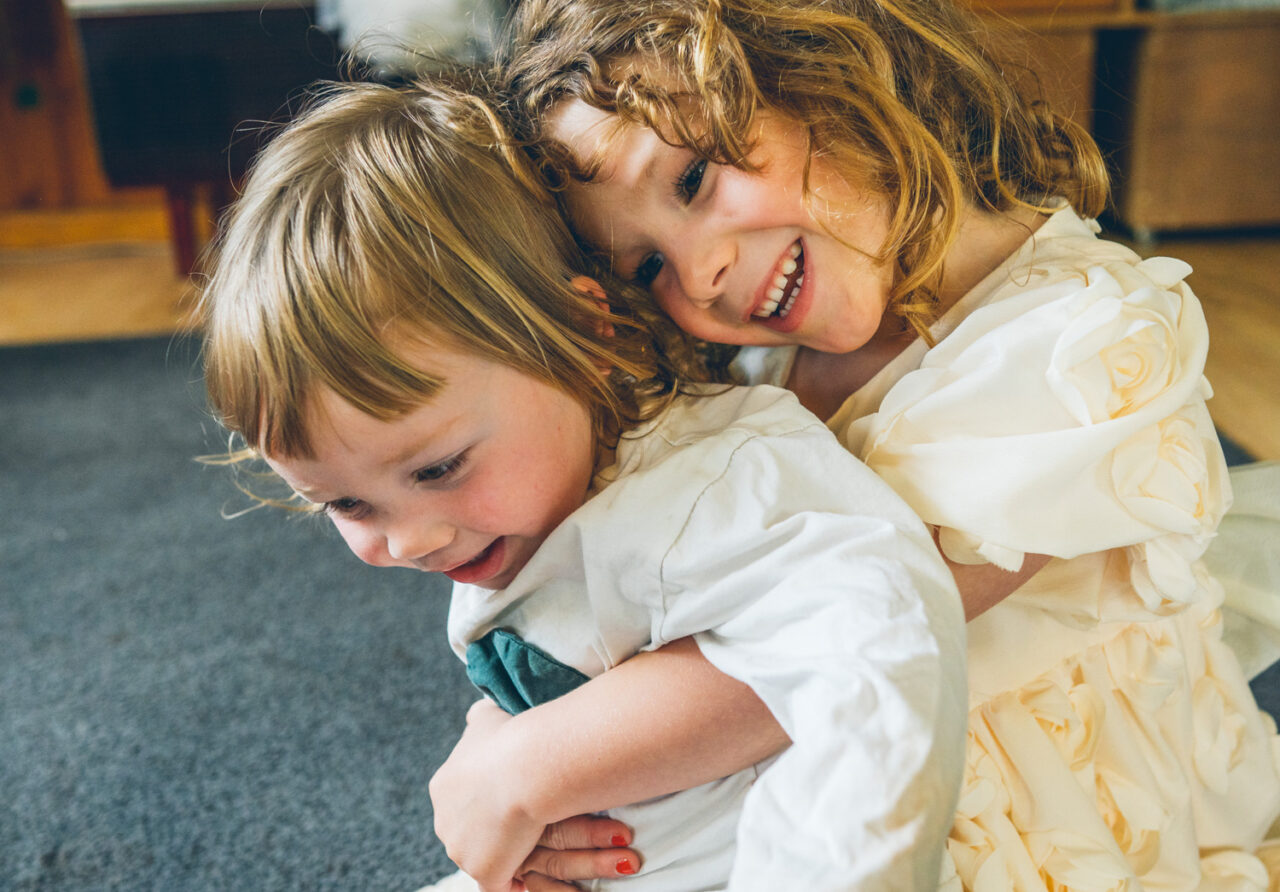Two young children in white dresses hugging and laughing together on the floor