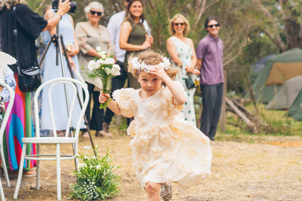 Young flower girl in a cream ruffled dress walking down the aisle holding a white bouquet and adjusting her flower crown, guests smiling behind her