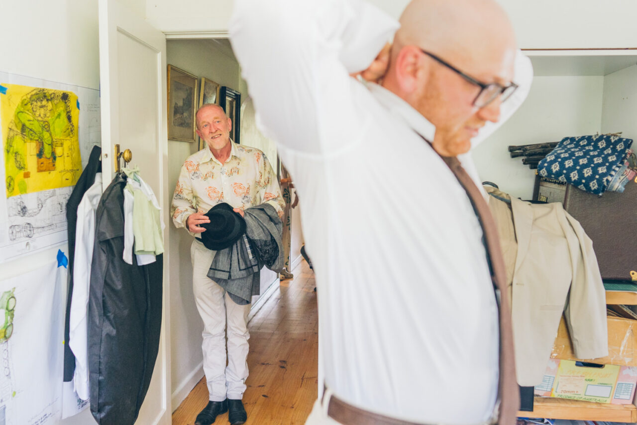 Dad looking on as his son is getting dressed. Together in a room, one smiling as he walks through a doorway carrying a hat and jacket
