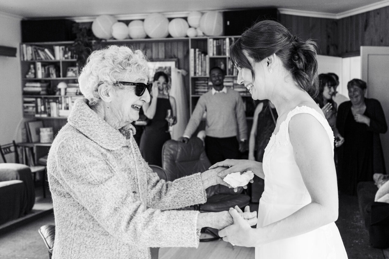 Black and white image of an elderly grandmother in sunglasses holding hands with a bride wearing her vintage wedding dress, surrounded by emotional family in the background