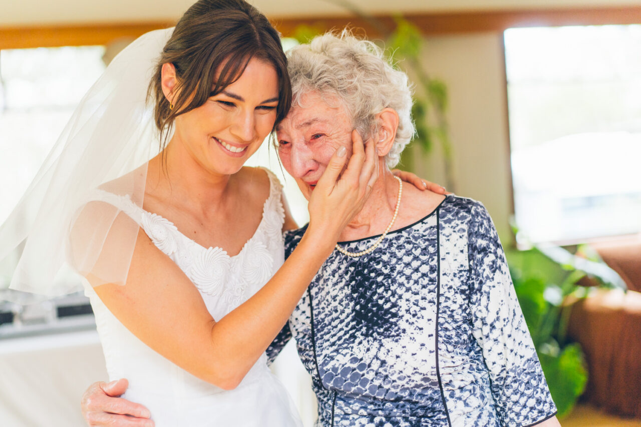 Bride in a vintage white wedding dress pressing her cheek against her grandmother's as the elderly woman wipes away tears with her hand