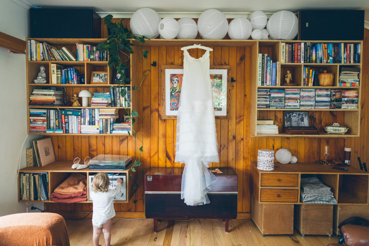 A vintage wedding dress hanging from a bookshelf in a warm timber-lined family home, with a small child standing below it