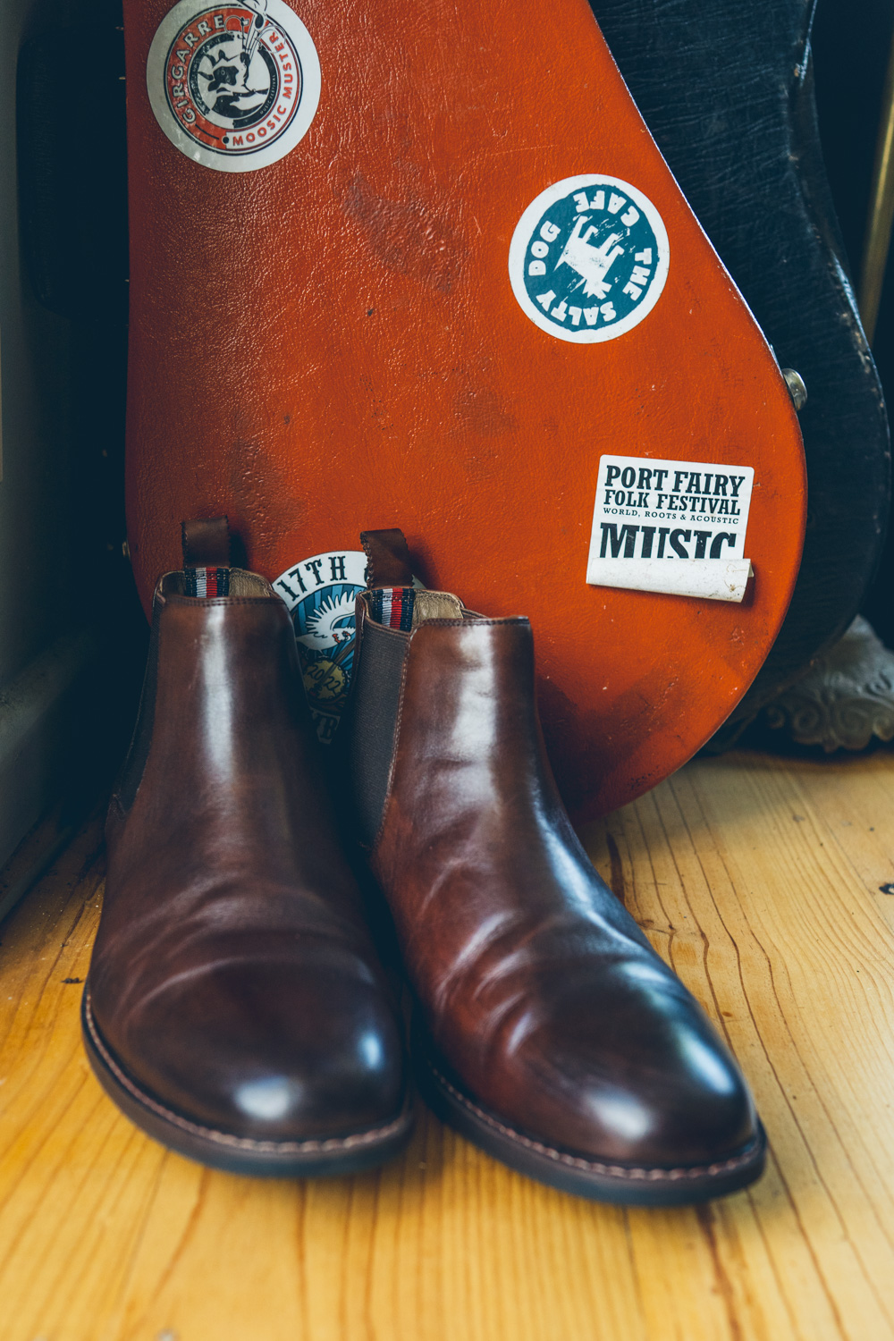Polished brown leather Chelsea boots resting against a stickered guitar case, including a Port Fairy Folk Festival sticker Caption: The details that tell you exactly who these people are.