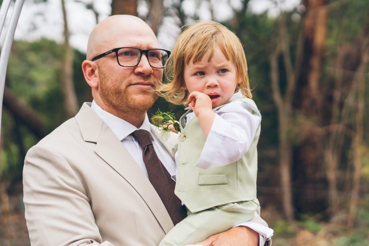 Groom in a beige suit holding a small child in a sage green vest at a backyard wedding ceremony on the Surf Coast