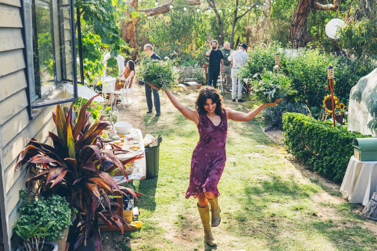 A woman in a purple dress and gumboots walking joyfully through a lush backyard garden holding flower arrangements above her head during a wedding reception
