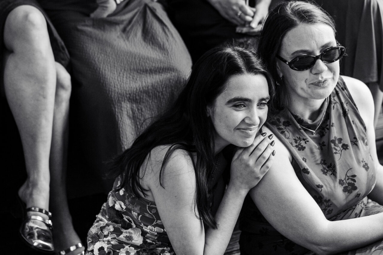 Black and white image of two women leaning into each other at a wedding, one smiling softly with tears in her eyes