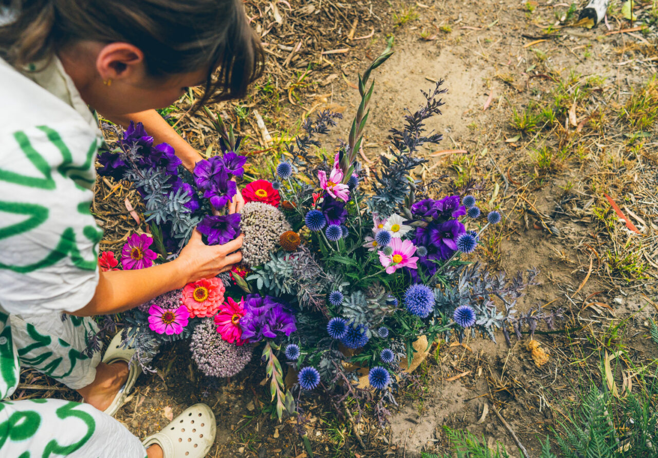 Person crouching over a colourful bunch of homegrown flowers including zinnias, globe thistles and cosmos, arranging them on the ground Caption: Every flower grown by hand. Every detail meaning something