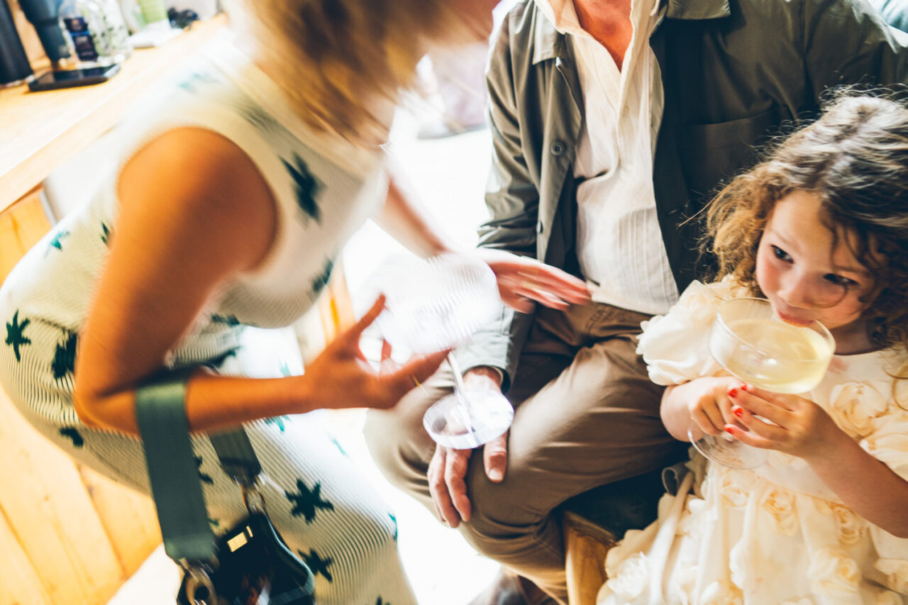 Young girl in a cream ruffled dress sipping from a cocktail glass while an adult talks animatedly beside her at a backyard wedding reception