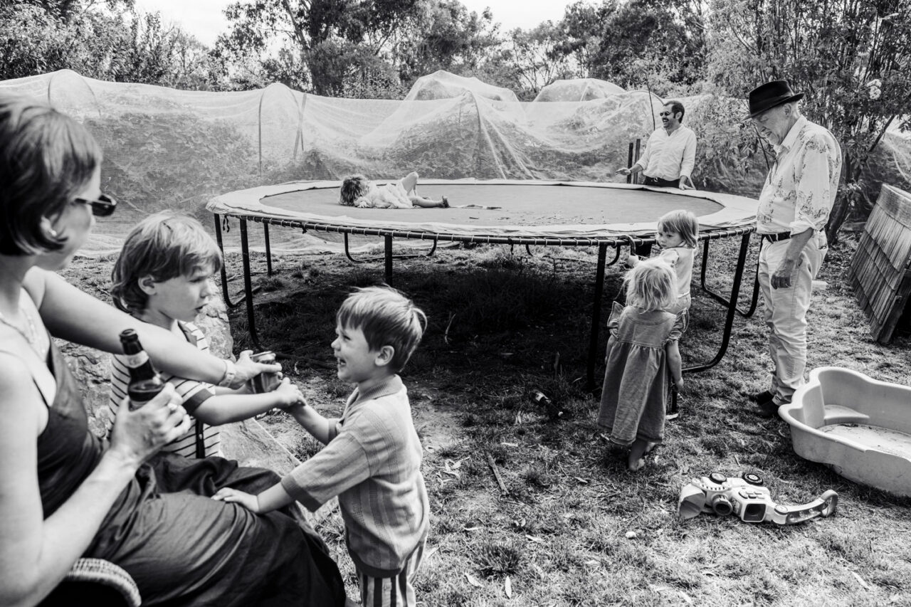 Black and white image of children playing on a trampoline in a backyard during a wedding reception while adults watch and socialise nearby