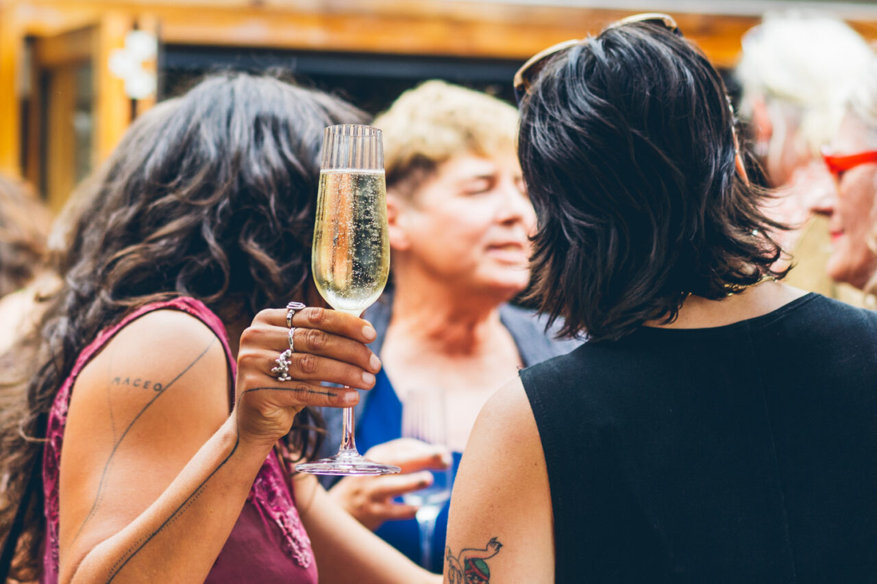 Two women holding champagne glasses and talking closely at a backyard wedding reception, fairy lights and guests in the background