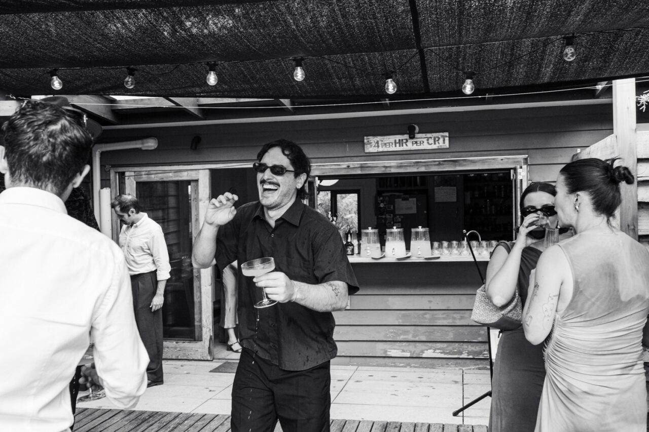 Black and white image of a guest laughing and holding a cocktail glass near the makeshift bar at a backyard wedding reception