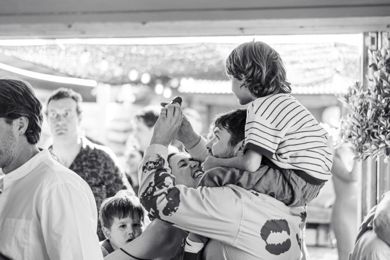 Black and white image of a man lifting a child above his head at a lively backyard wedding reception while another small child watches from below
