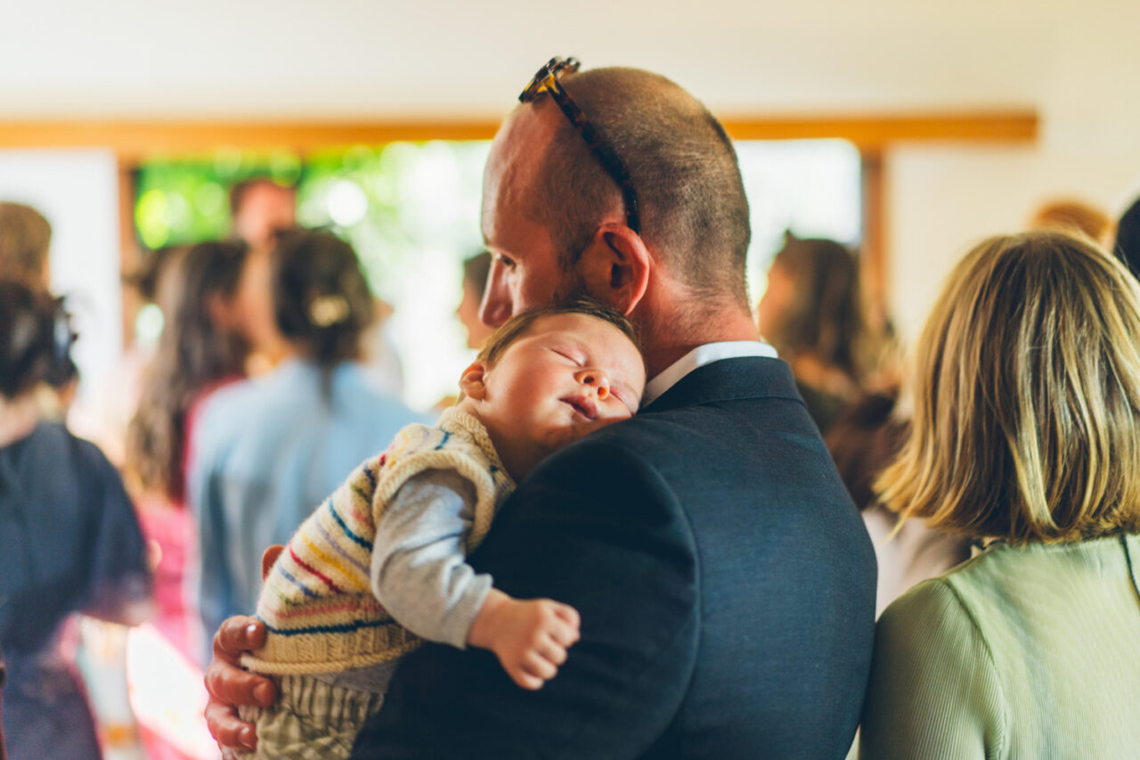 A baby in a striped knit jumper sleeping soundly on a guest's shoulder at a backyard wedding reception