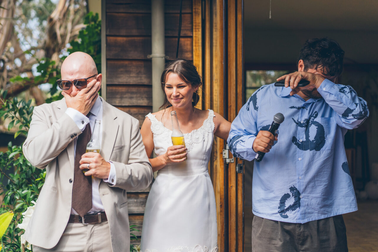 Groom wiping tears during speeches at a backyard wedding reception while the bride stands beside him smiling and a friend holds a microphone