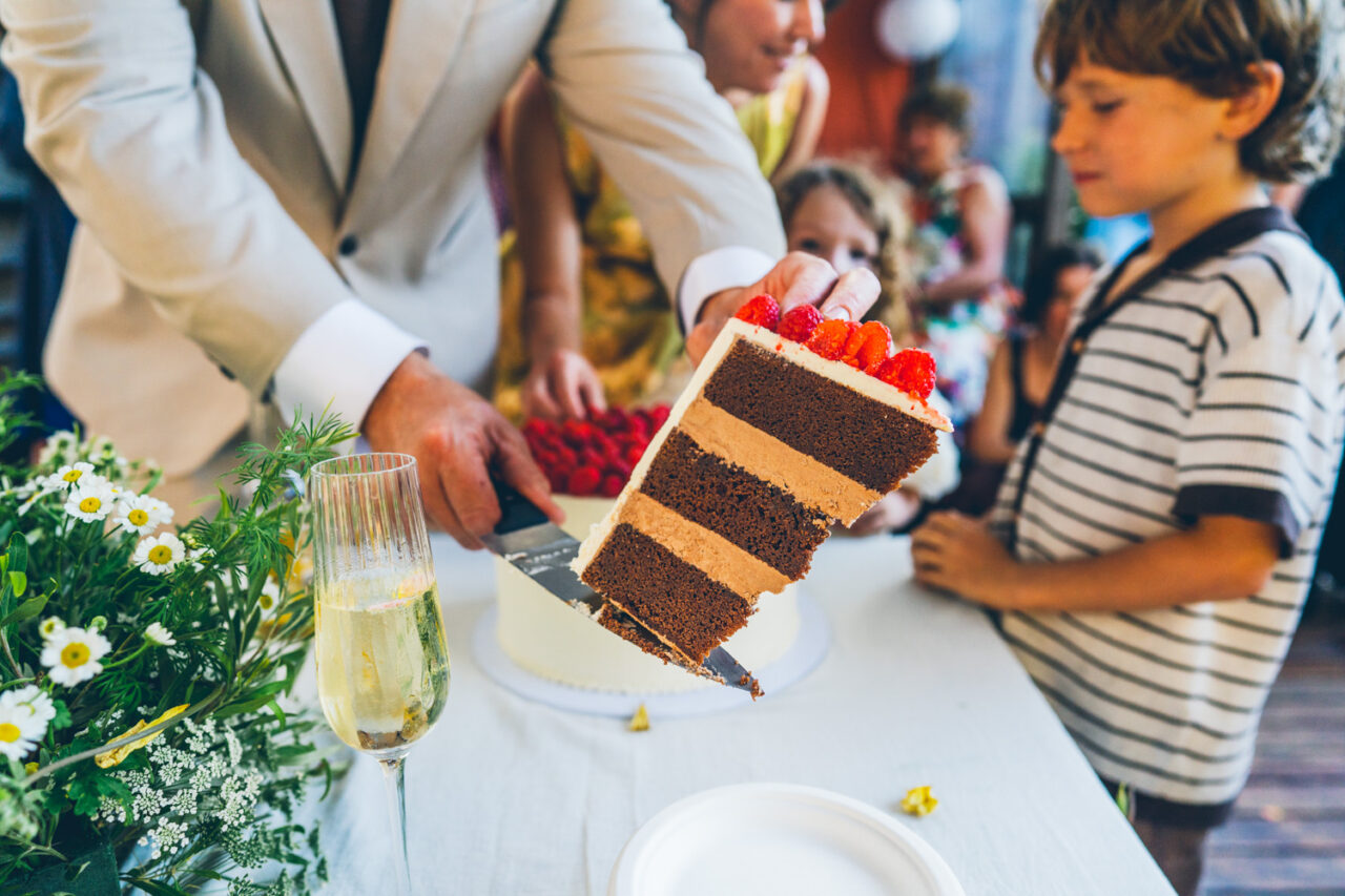 Groom lifting a large slice of layered chocolate wedding cake from Ginger and Spice while a child watches eagerly beside homegrown flower arrangements