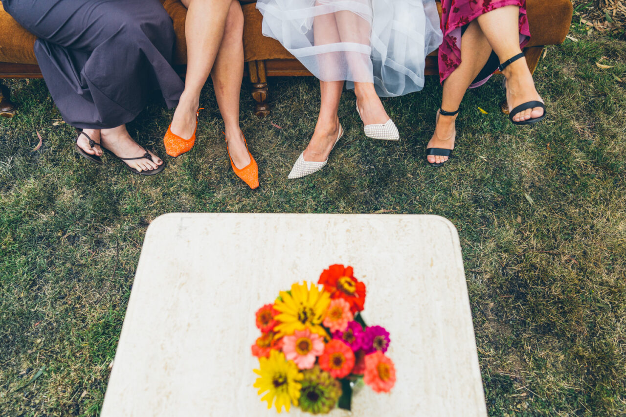 Overhead shot of four pairs of shoes worn by women sitting on a couch at a backyard wedding, with homegrown flowers in a vase on a table below