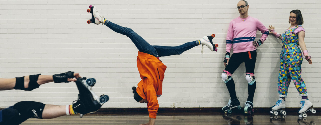 A roller skater does a handstand in the middle of a rink while two others in colourful retro outfits lean against a white brick wall watching, and a fourth skater's legs appear in the foreground, captured during a brand documentary session for YMCA CoMA by LJM Photography