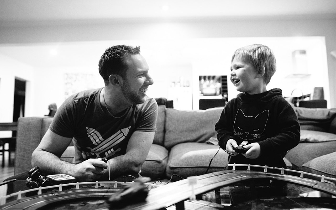 A father and young child laugh together over a slot car racing track on the living room floor, captured in black and white during an in-home Your Story documentary session by LJM Photography