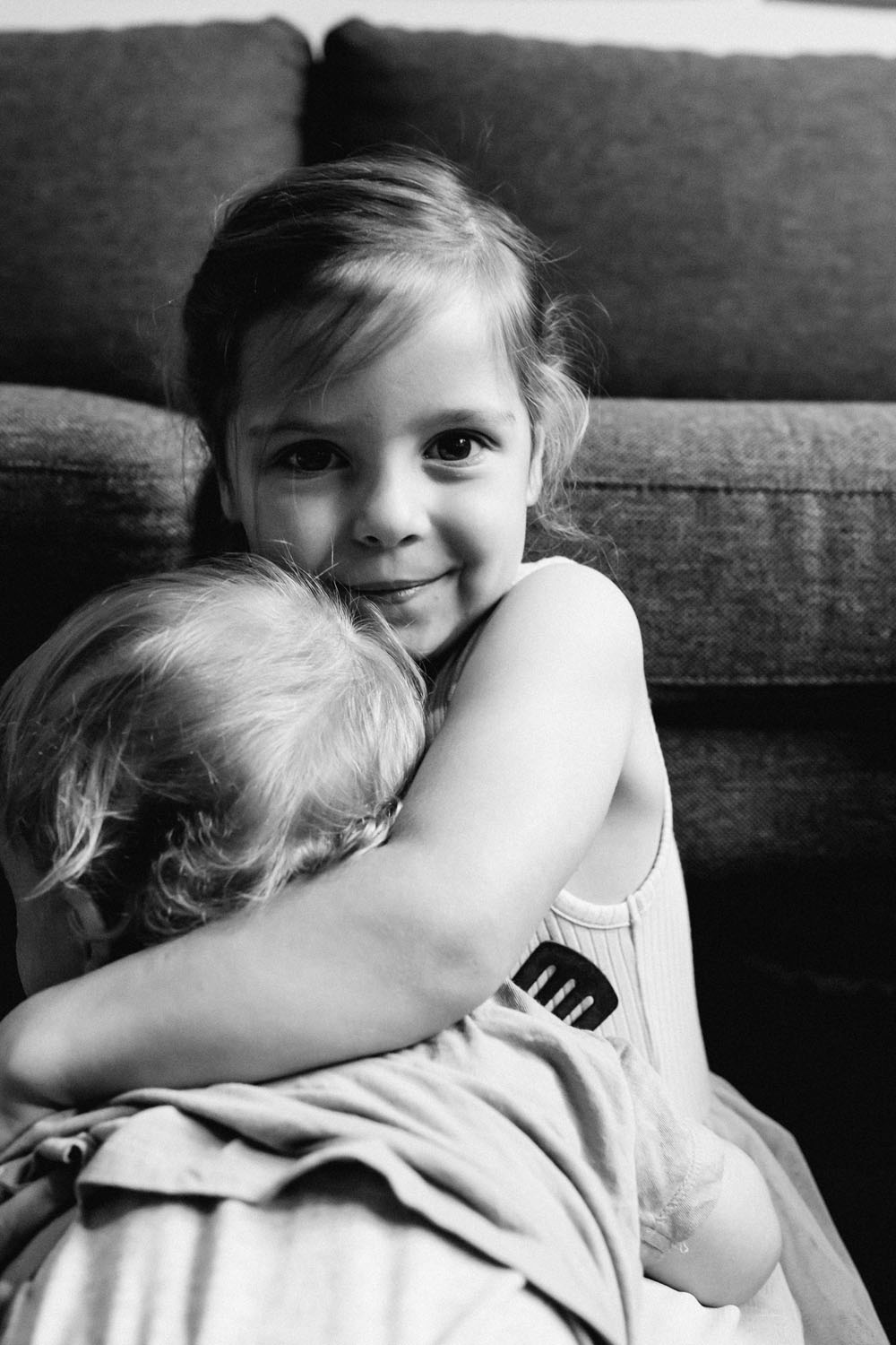 Black and white documentary portrait of a young girl hugging a toddler sibling and smiling directly at the camera, Your Story family session by LJM Photography Surf Coast Melbourne
