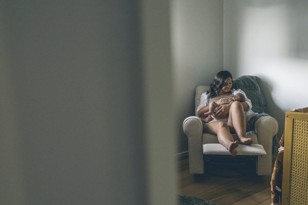 A mother sits in a nursing chair in soft natural light, breastfeeding her newborn in the corner of a quiet nursery, photographed through a doorway during an in-home documentary family session