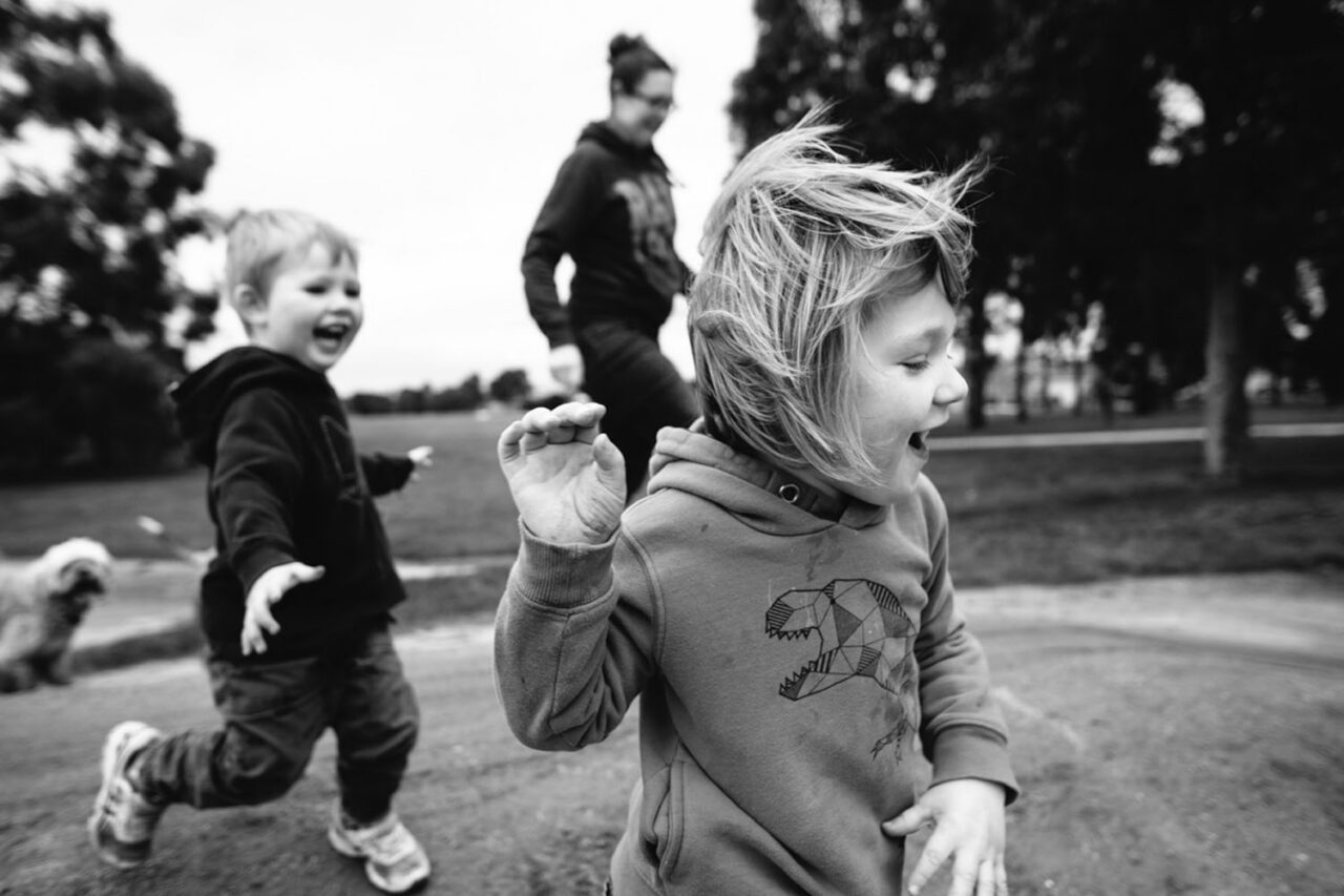 Two young children running and laughing in a park with a parent in the background, black and white documentary family photography by LJM Photography