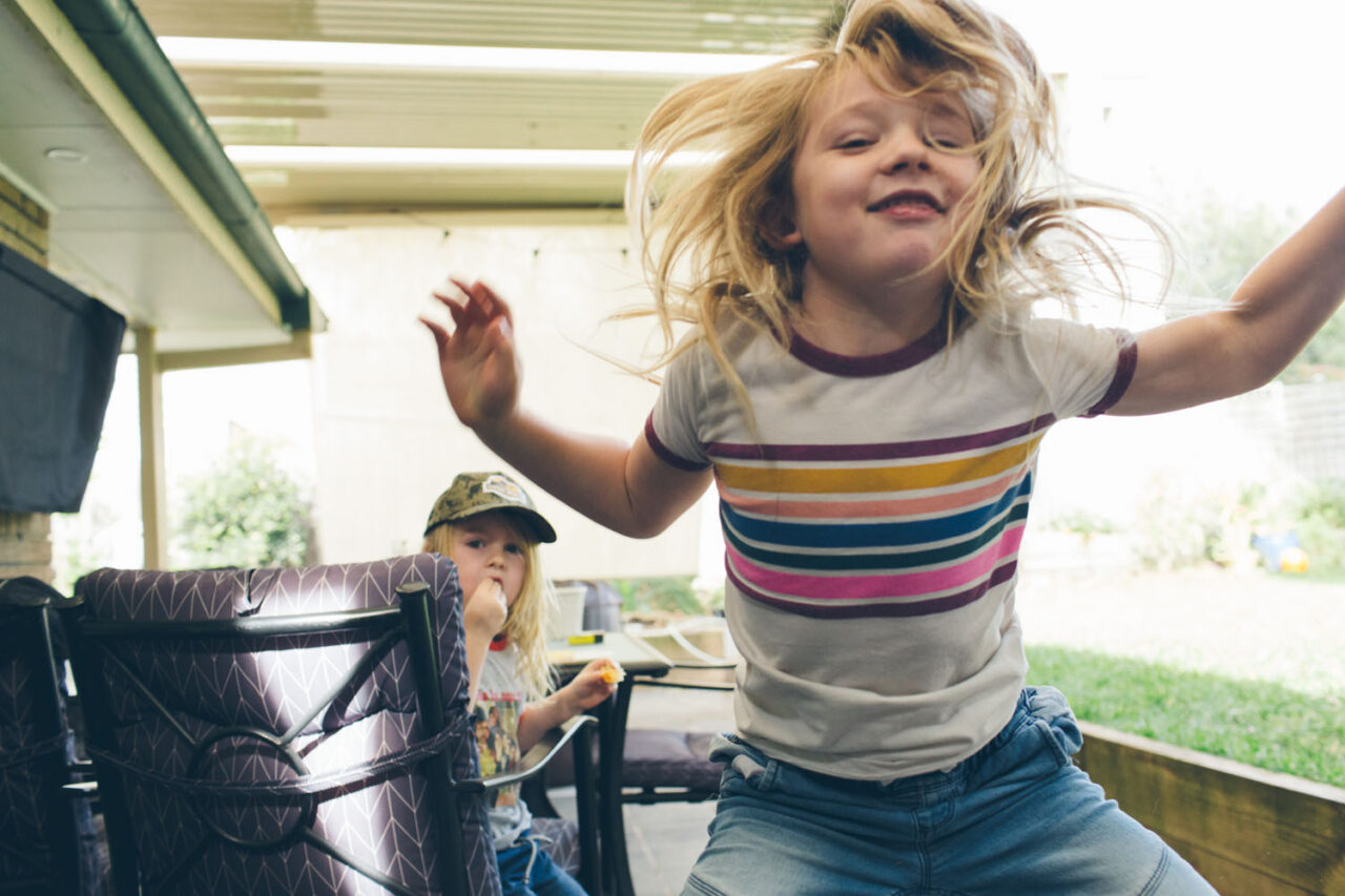 A young boy jumping with arms outstretched in a backyard while a sibling watches, documentary family photography by LJM Photography