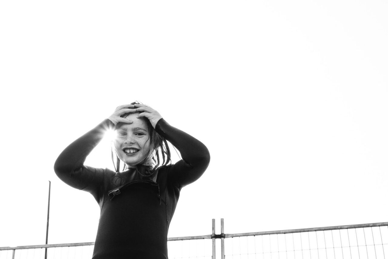 A child in a wetsuit grinning with hands on head, sunlight flaring through fingers, black and white documentary photography by LJM Photography