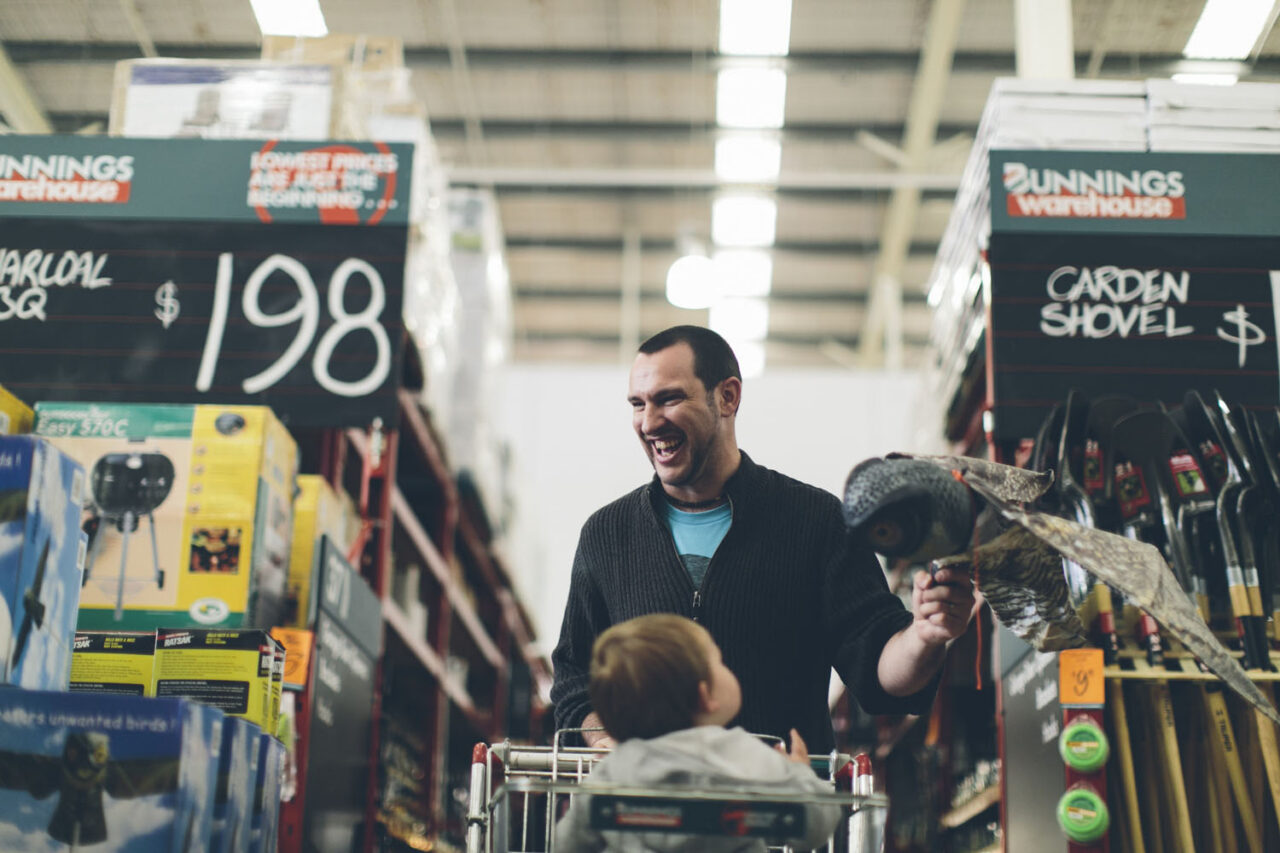 A father laughing while pushing a baby in a trolley through a Bunnings warehouse, documentary family photography by LJM Photography