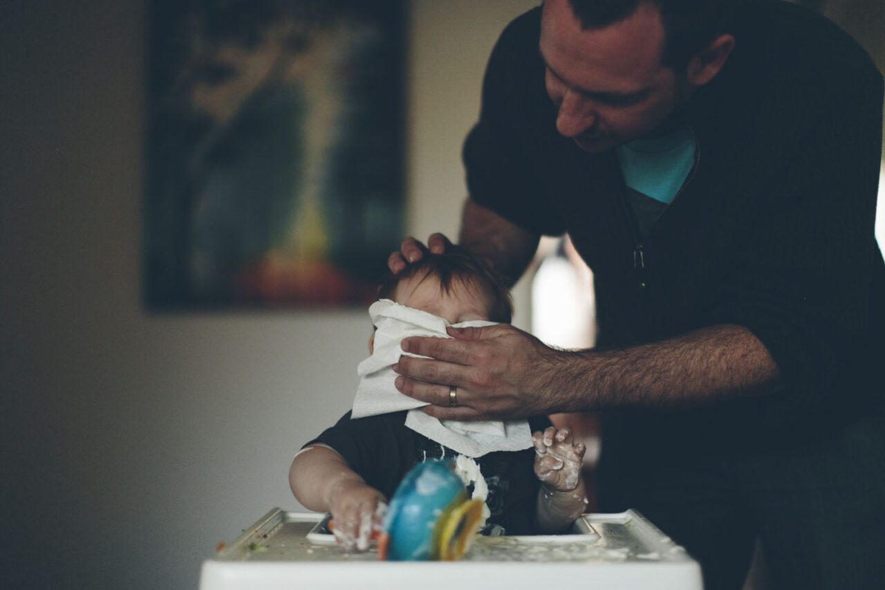 A father gently wiping a baby's face at a highchair, documentary family photography by LJM Photography