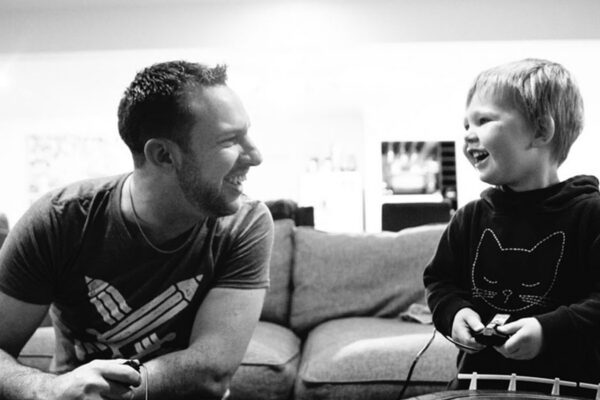 A father and young son laughing together on the couch playing video games, captured in black and white by LJM Photography