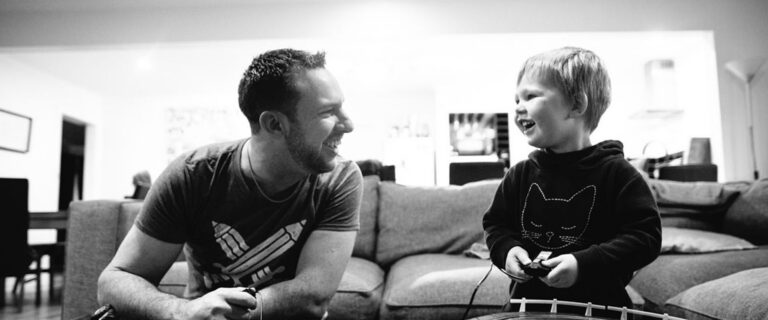 A father and young son laughing together on the couch playing video games, captured in black and white by LJM Photography