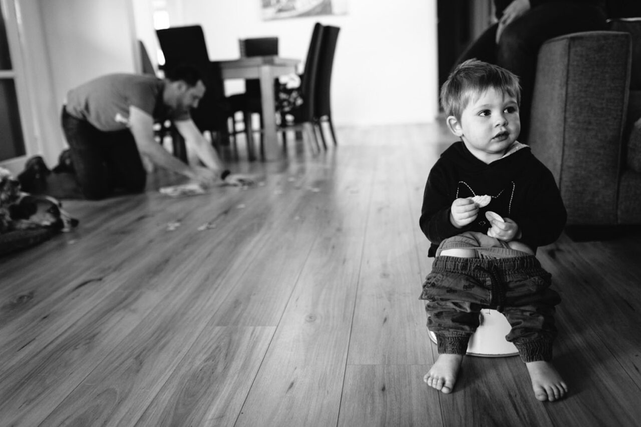 A toddler sitting on a potty eating a snack while a father cleans up mess in the background, black and white documentary family photography by LJM Photography
