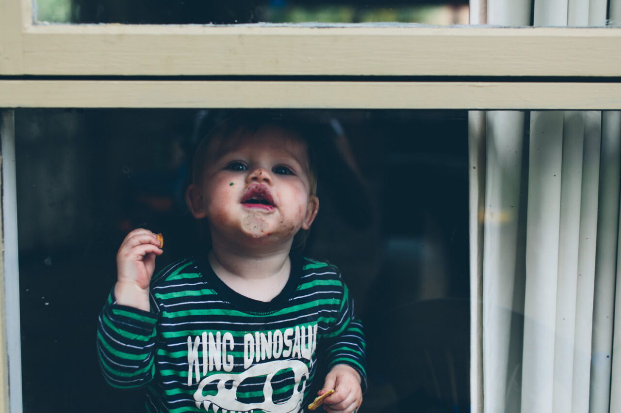 A toddler with a messy face standing at a window wearing a King Dinosaur top, documentary family photography by LJM Photography