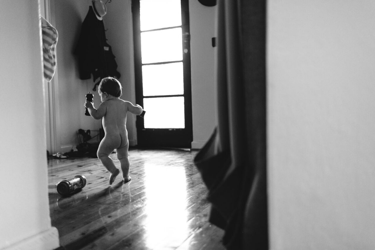 A toddler running naked down a sunlit hallway holding an object, captured in black and white by LJM Photography