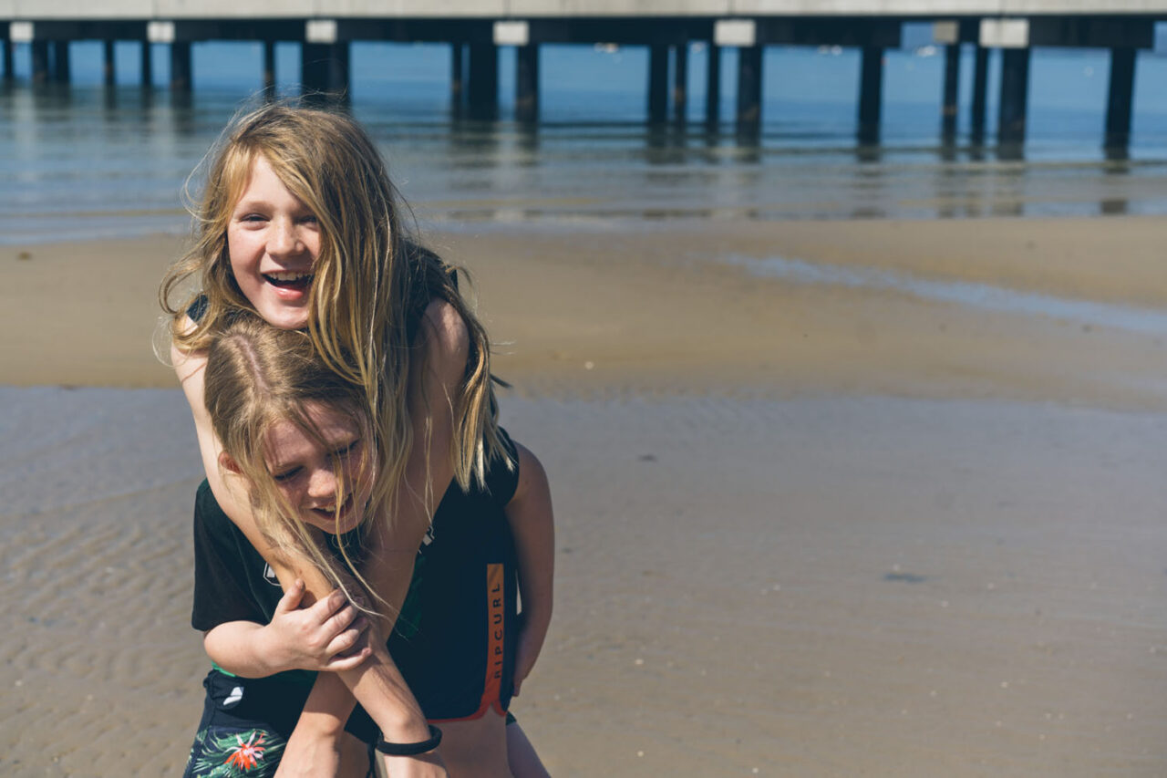 Two children at the beach, one giving the other a piggyback and laughing, pier in the background, documentary family photography by LJM Photography