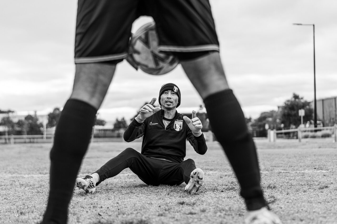 AUFC goalie sitting on the ground gesturing playfully, framed between two players' legs, black and white