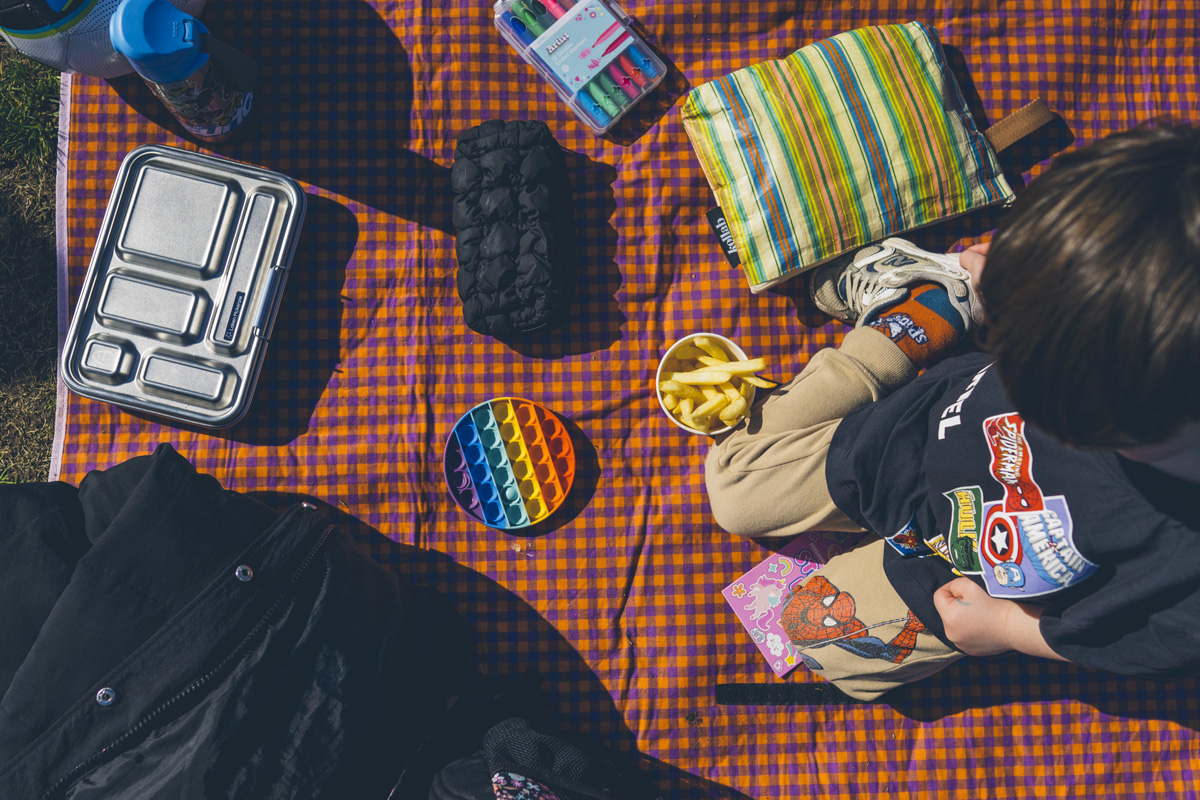 Overhead shot of a picnic rug with chips, toys and a young sibling at a soccer match