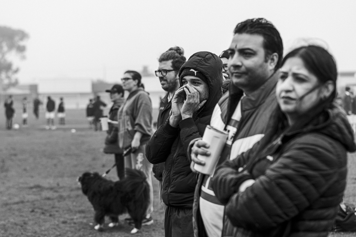 Parents watching a junior soccer match from the sideline in fog, one with a dog