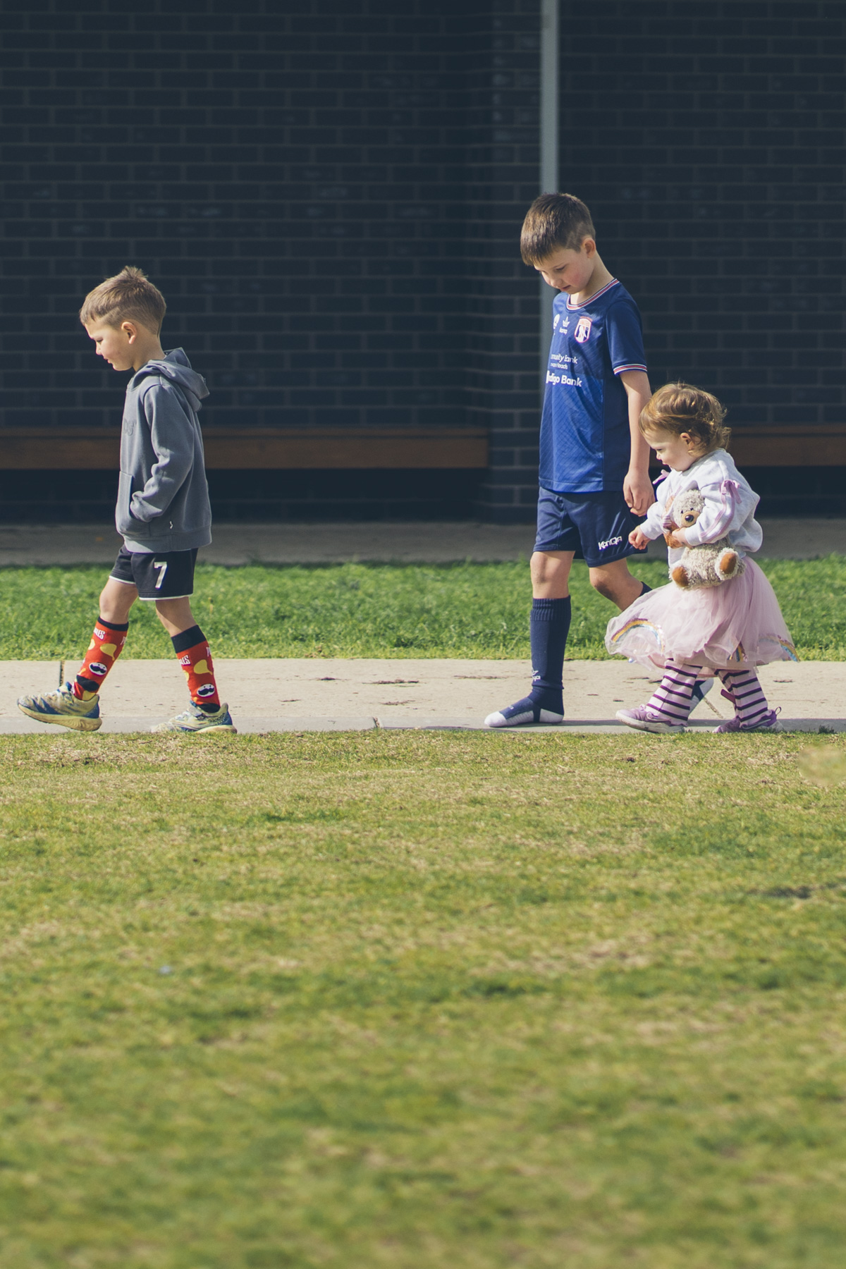 hree siblings walking together, one in soccer kit and one in a tutu carrying a teddy bear